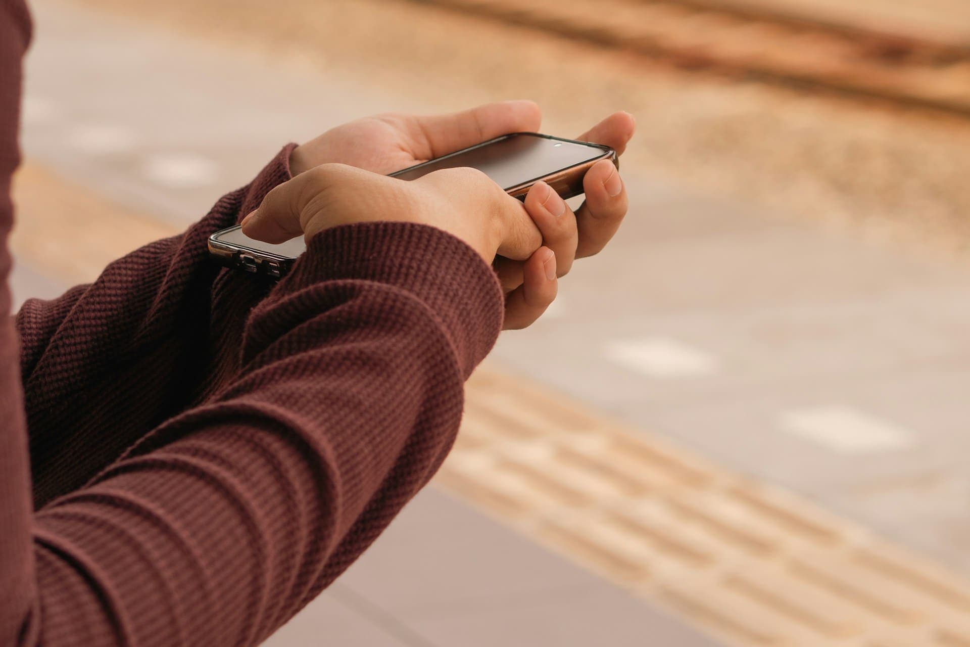A person in a maroon knitted sweater holding a smartphone in both hands, with a blurred paved walkway behind